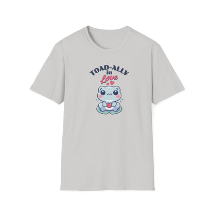 a cartoonish blue frog with a cheerful expression holds a small red heart in its hands. The design is printed on a ice grey T-shirt and features the playful phrase 'Leap into Love!' adding a charming and romantic vibe.