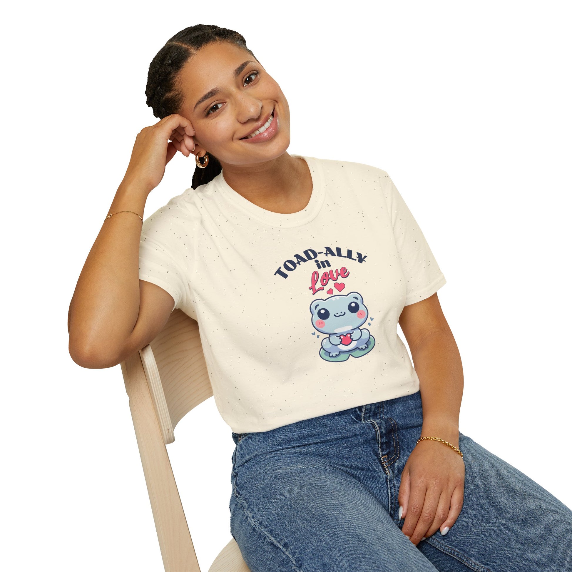 a woman wearing a cartoonish blue frog with a cheerful expression holds a small red heart in its hands. The design is printed on a natural colored T-shirt and features the playful phrase 'Leap into Love!' adding a charming and romantic vibe.