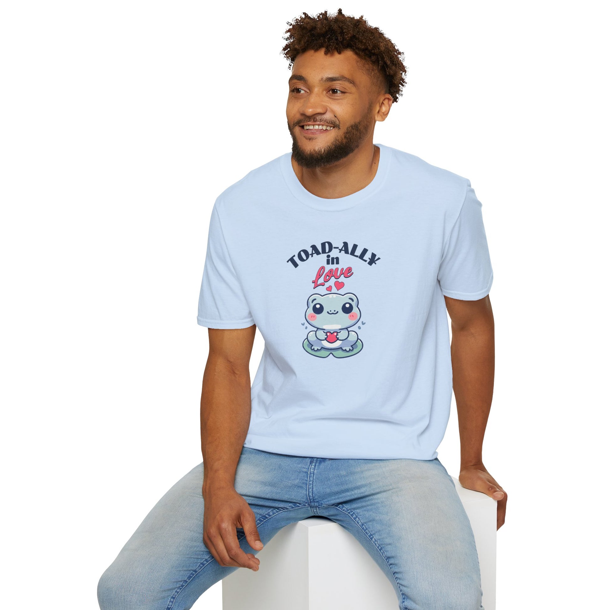 a man wearing wearing a cartoonish blue frog with a cheerful expression holds a small red heart in its hands. The design is printed on a light blue T-shirt and features the playful phrase 'Leap into Love!' adding a charming and romantic vibe.