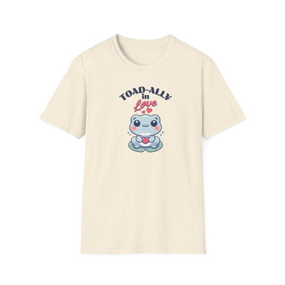 a cartoonish blue frog with a cheerful expression holds a small red heart in its hands. The design is printed on a natural colored T-shirt and features the playful phrase 'Leap into Love!' adding a charming and romantic vibe.